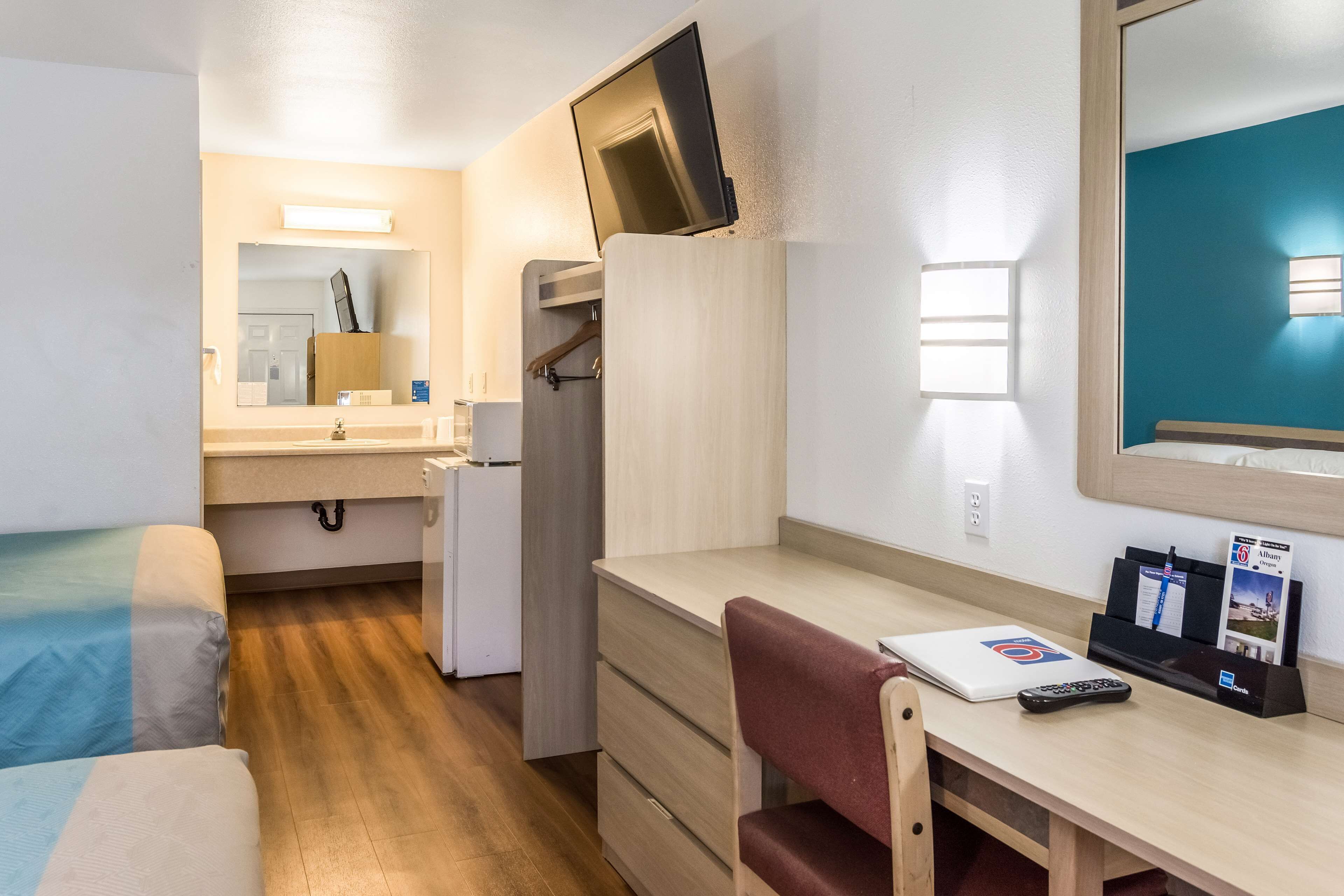 A compact hotel room with a bed, a desk with a laptop, a chair, a TV mounted on the wall, and a small refrigerator.