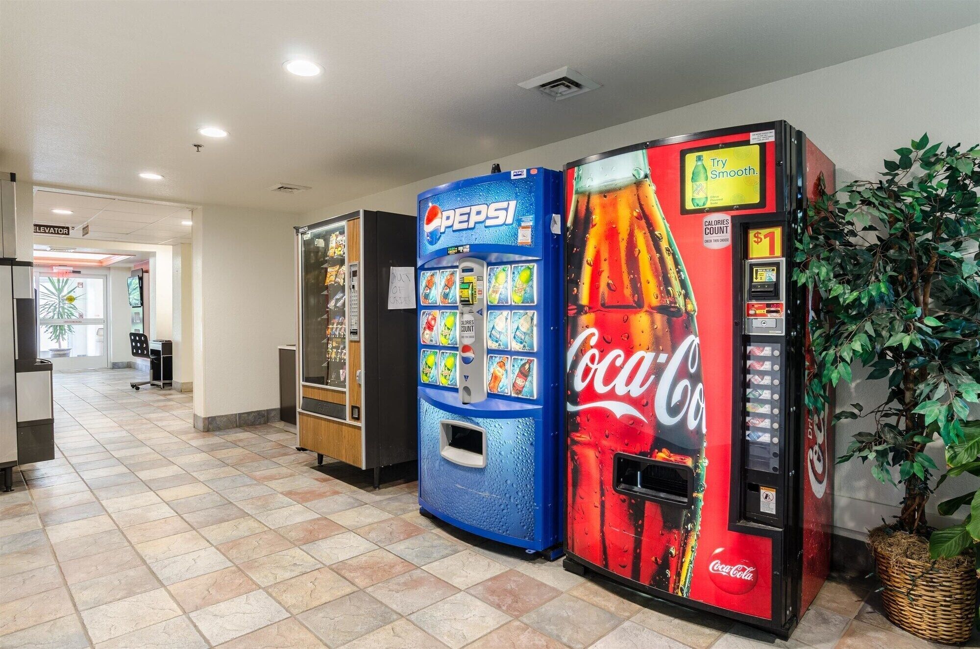 A vending area with Pepsi and Coca-Cola machines, a plant, and a sign for an elevator.