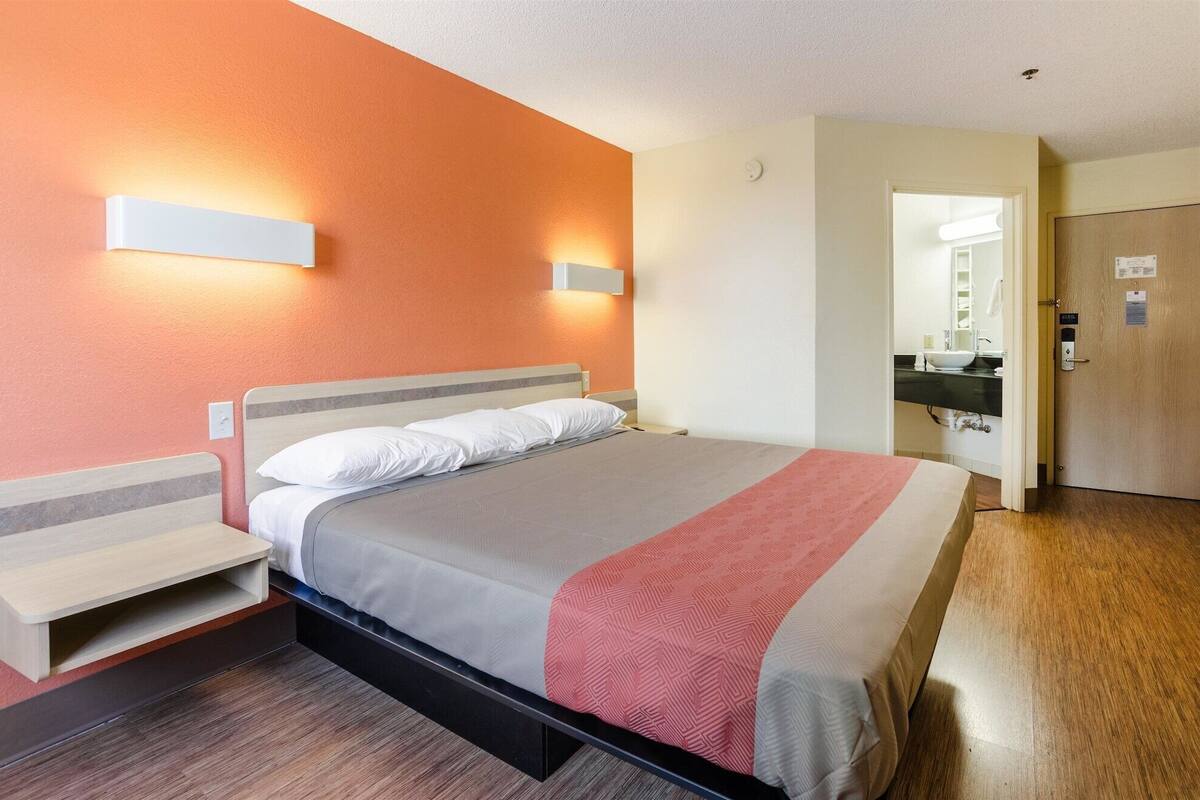A hotel room with a bed, bedside tables, a nightstand, a wooden floor, a bathroom with a sink and mirror, and an orange wall.