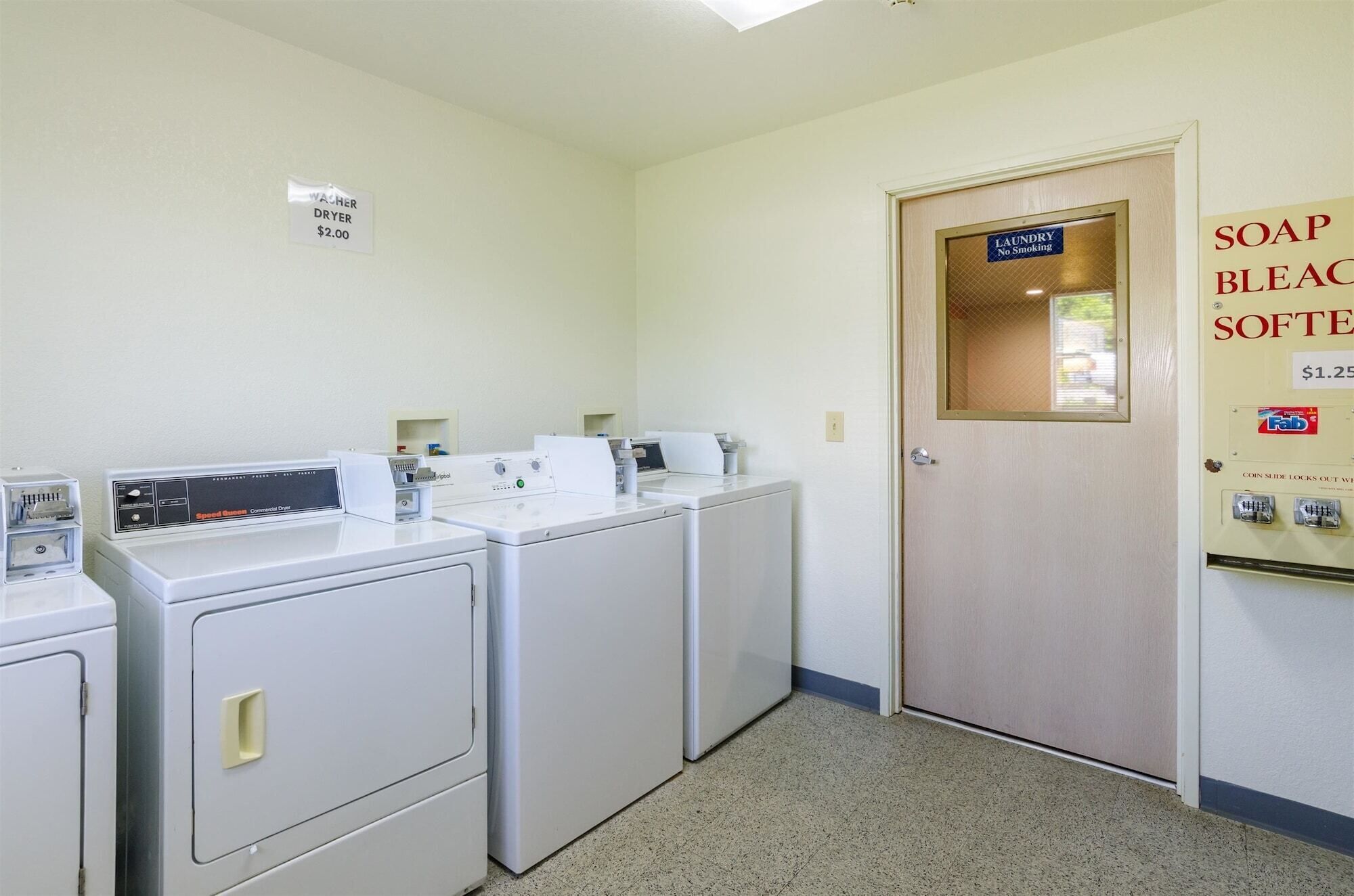 A laundry room with two front-loading washers and dryers, a sign indicating a $2.00 washer and dryer fee, a door with a sign for laundry and no smoking, and a soap and bleach softener dispenser with a price of $1.25.