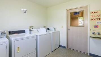 A laundry room with two front-loading washers and dryers, a sign indicating a $2.00 washer and dryer fee, a door with a sign for laundry and no smoking, and a soap and bleach softener dispenser with a price of $1.25.