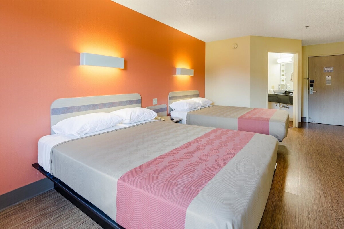 A hotel room with two beds, each with a headboard and bedside tables. There is a bathroom visible in the background with a sink and mirror.