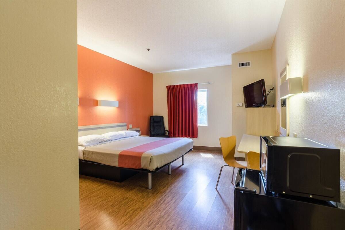A hotel room with a bed, a desk with a computer, a chair, a TV, and a window with curtains.