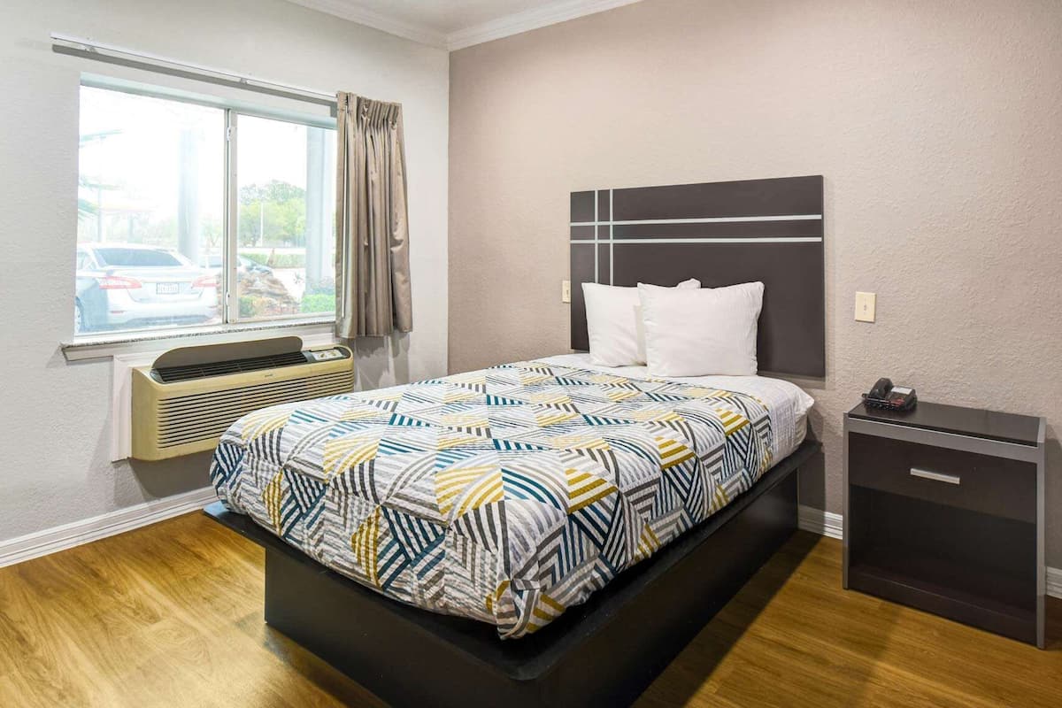 A bedroom with a bed, a bedside table, a nightstand, a window with curtains, and an air conditioning unit.
