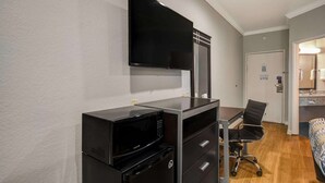 Une chambre d’hôtel compacte dotée d’une kitchenette, d’un téléviseur à écran plat fixé au mur, d’un petit bureau avec une chaise et d’une salle de bain visible à travers une porte ouverte.