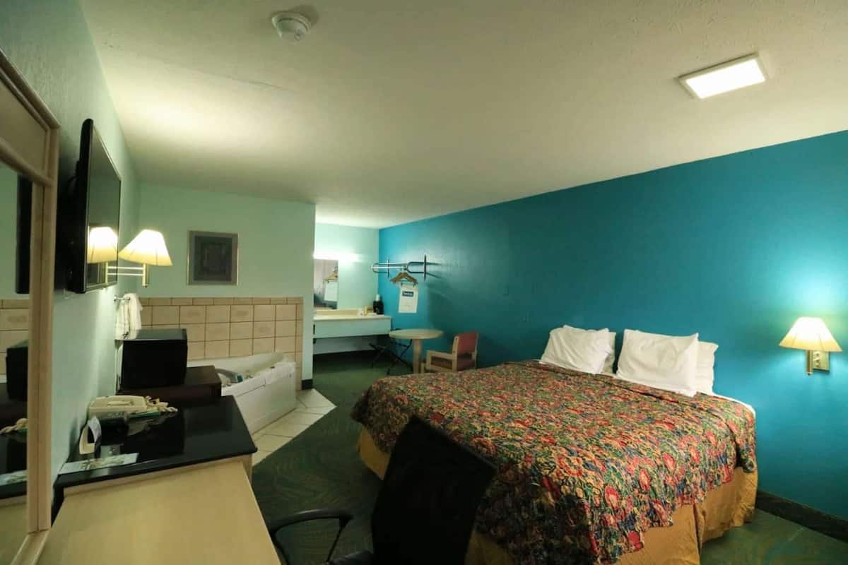 A hotel room with a bed, a desk, a chair, a mirror, and a wall-mounted lamp.