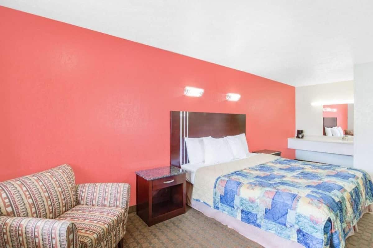 A hotel room with a bed, a nightstand, a chair, a mirror, and a red wall.