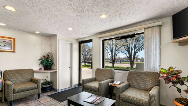 Eine Lobby mit einer Rezeption, zwei Sesseln, einem Couchtisch und einem großen Fenster mit Blick auf Bäume und einen Parkplatz.