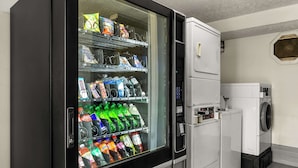 Ein Verkaufsautomat mit verschiedenen Snacks und Getränken, eine Waschmaschine und ein Trockner in einem Raum mit Fliesenboden.