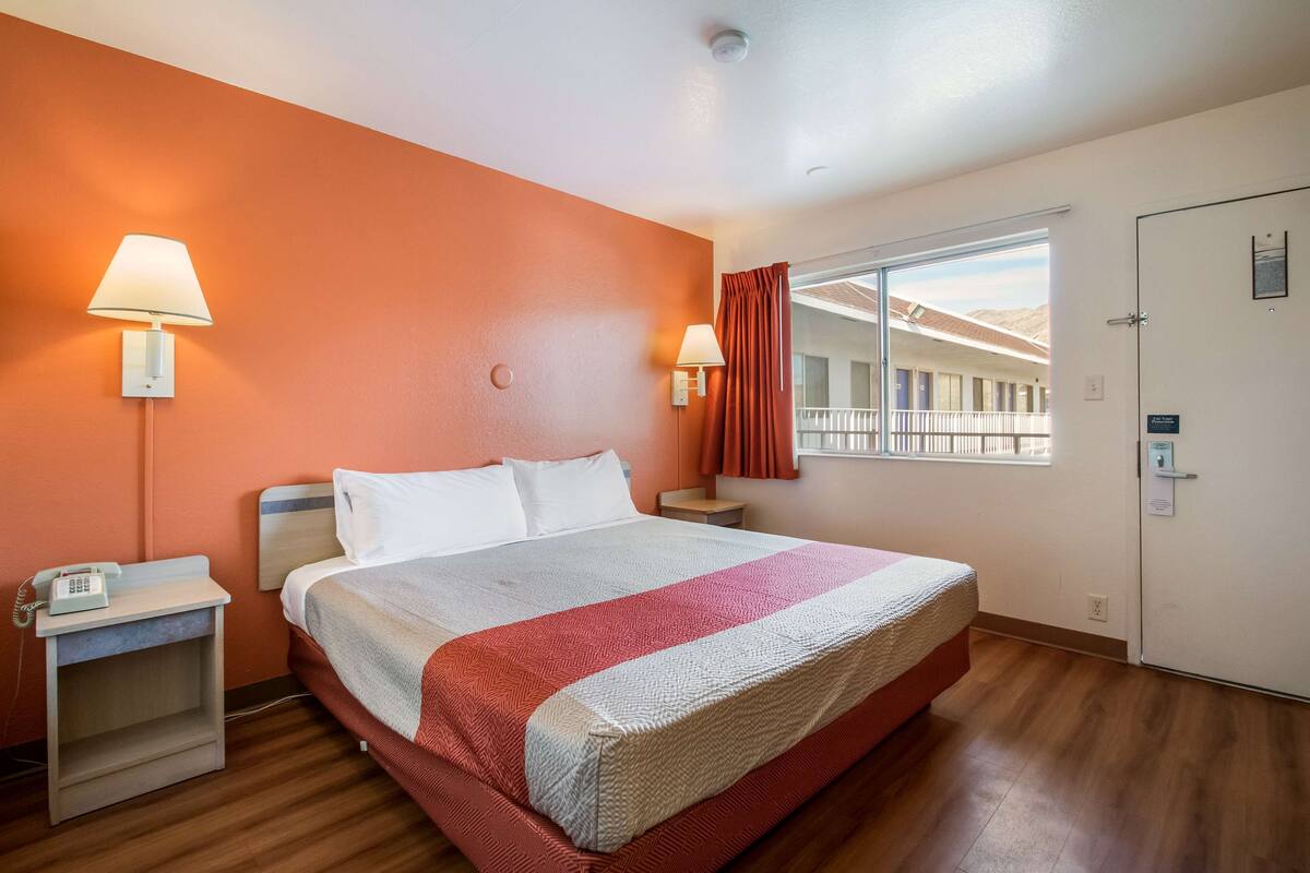 A hotel room with a large bed, two bedside lamps, a nightstand with a phone, a window with curtains, and a door with a sign.