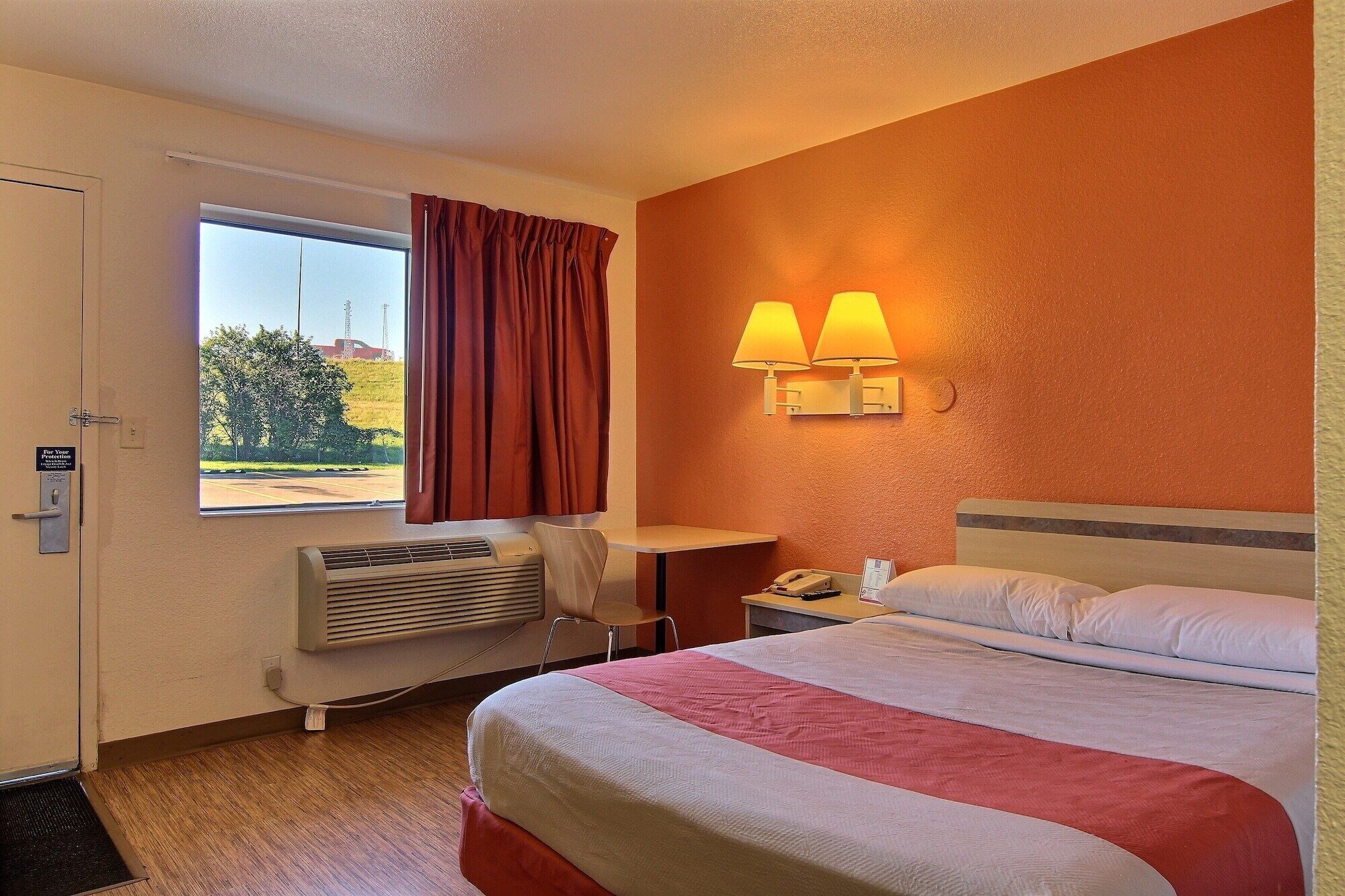 Una camera d'albergo con un letto, una scrivania, una sedia, una finestra con tende e due lampade a parete.