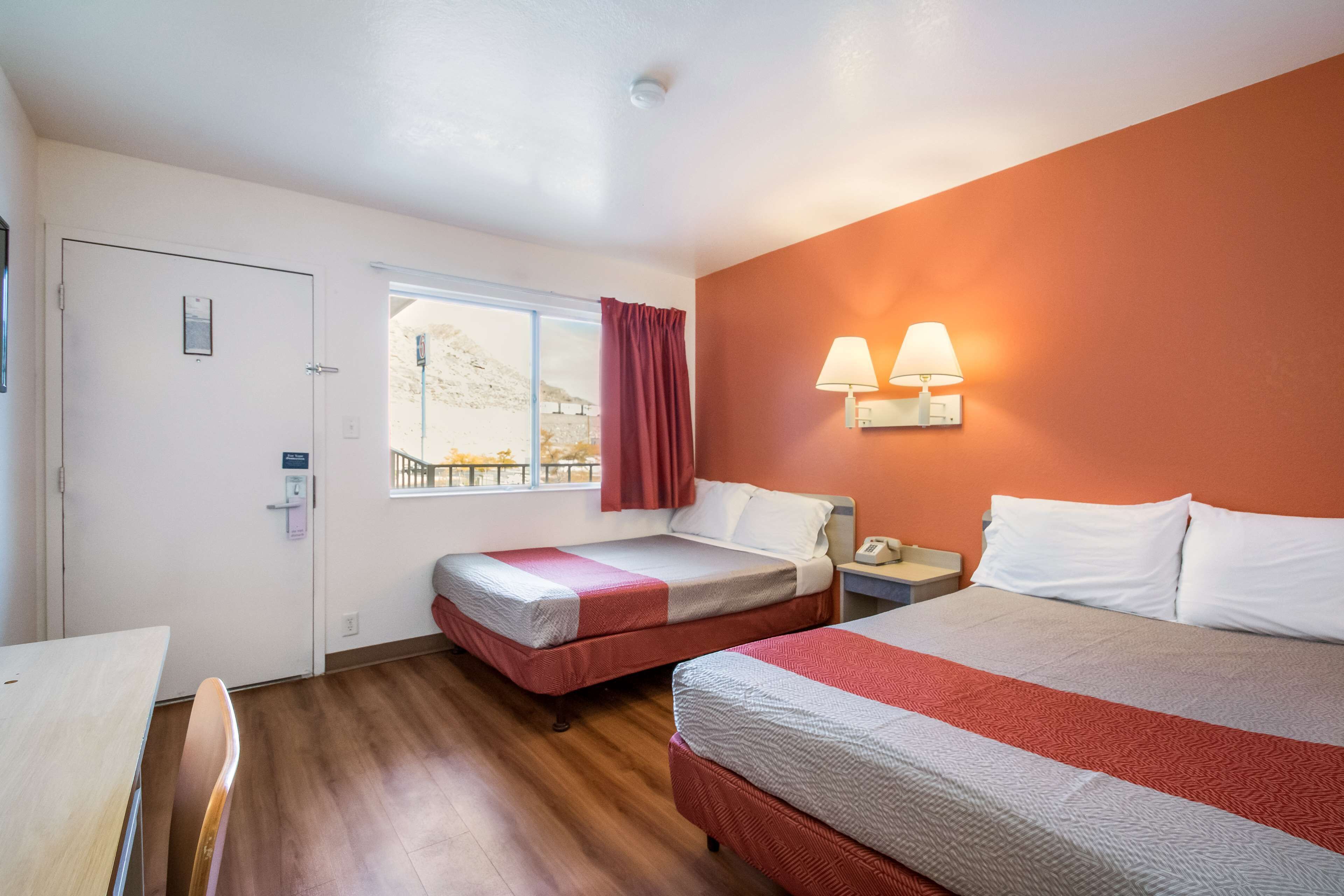 A hotel room with two beds, a desk, a window with a view, and a wall-mounted lamp.