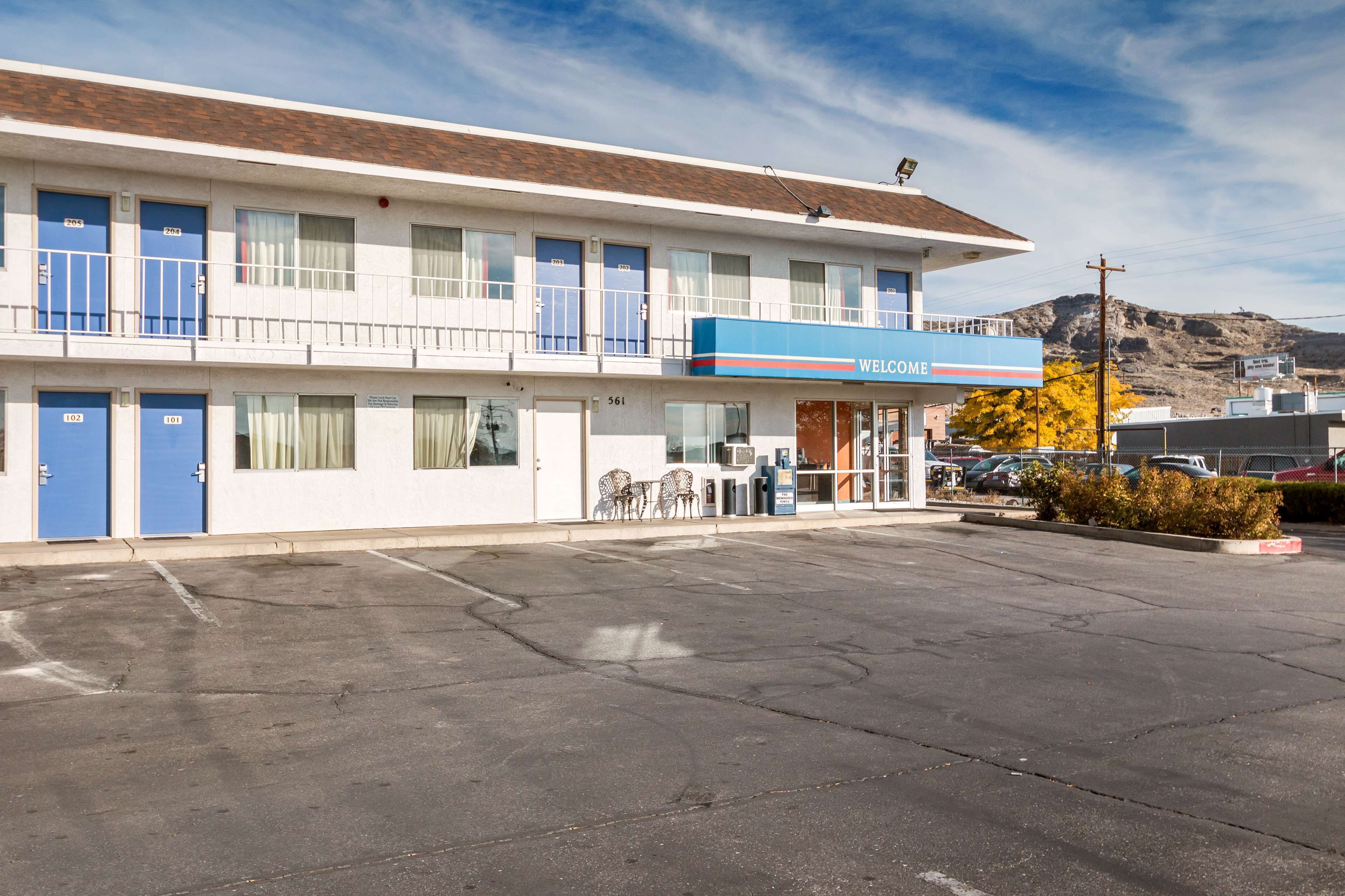 A motel with a blue 'Welcome' sign, multiple rooms, a parking lot, and a mountain in the background.