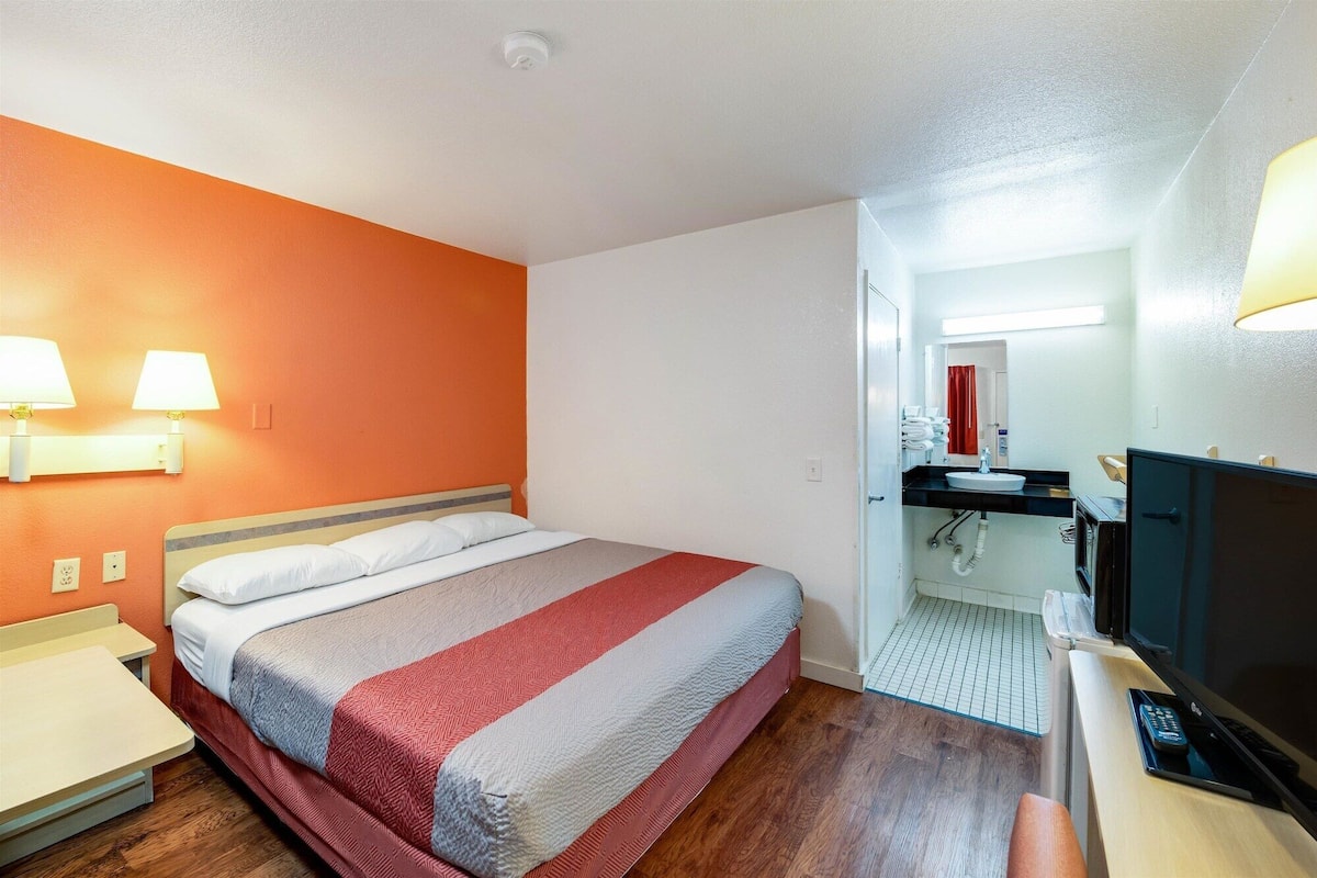 Een hotelkamer met een bed, nachtkastjes, een televisie, een badkamer met een wastafel en spiegel, en een rood gordijn.