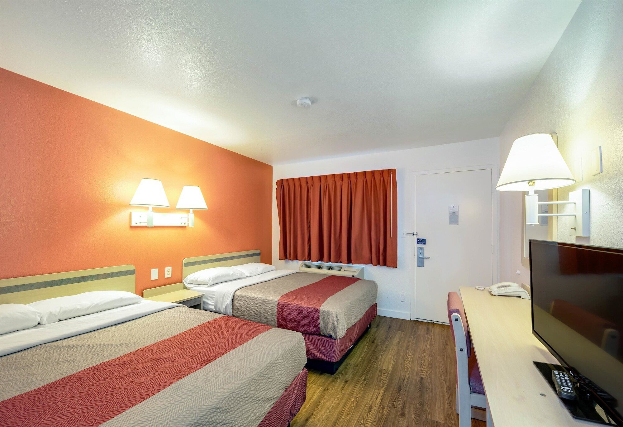 A hotel room with two beds, a desk with a TV and phone, and a door.