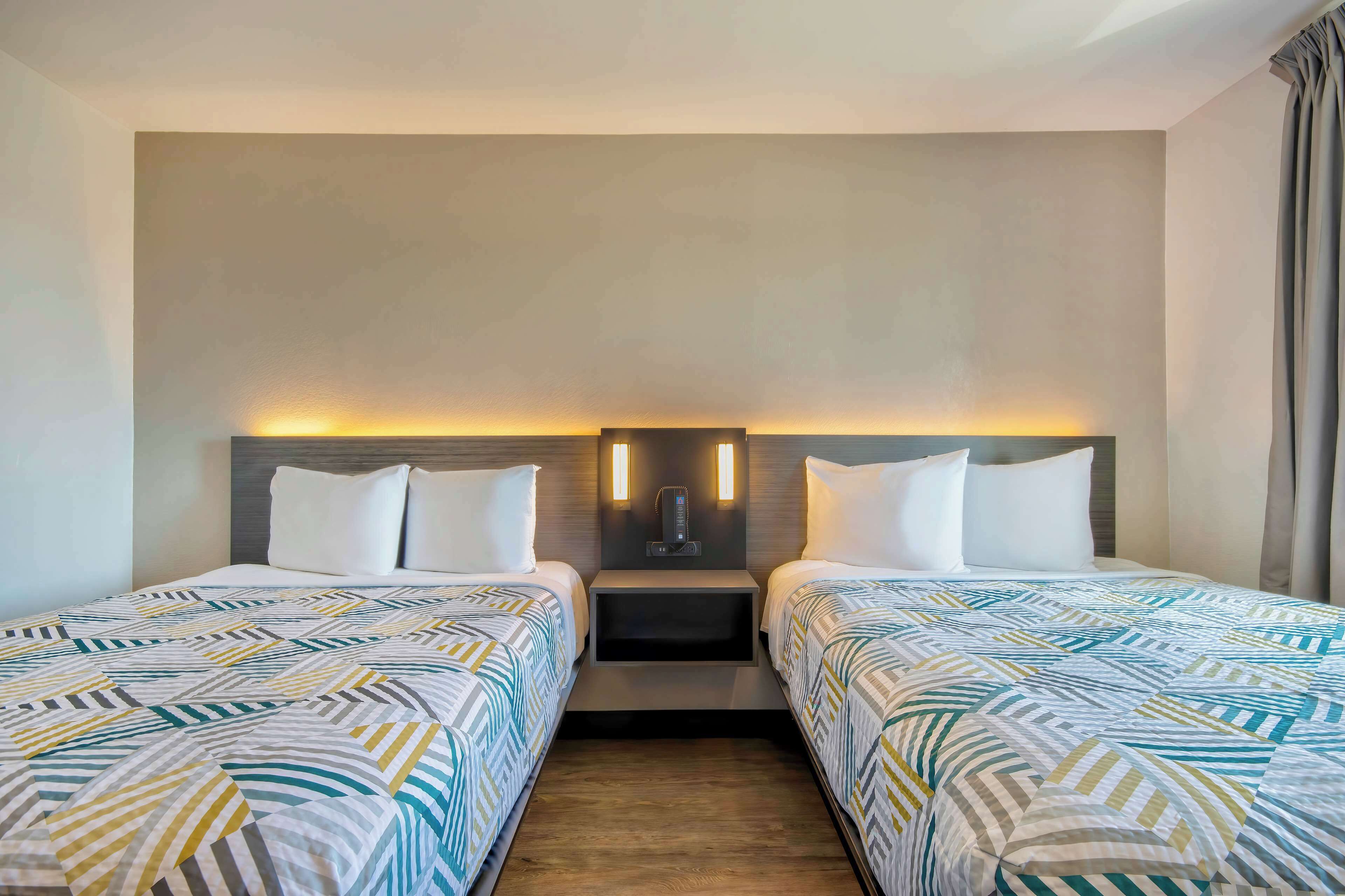 A hotel room with two beds, a nightstand with a lamp, and a wall-mounted light fixture.