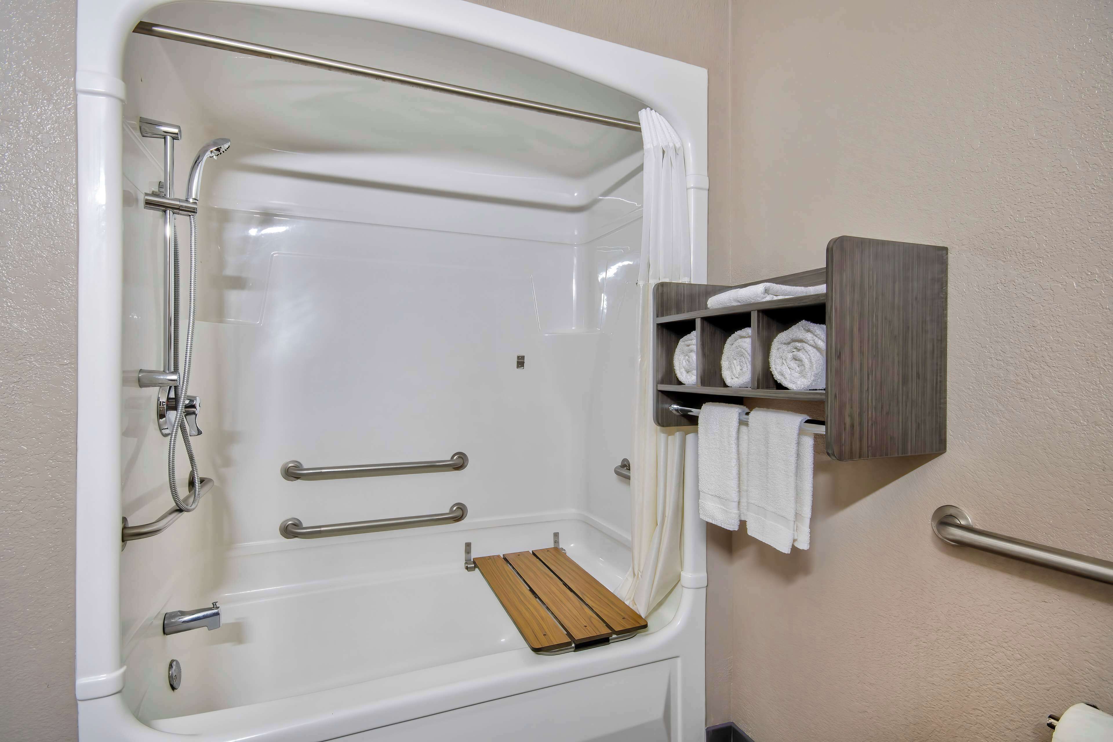 A bathtub with a wooden seat, grab bars, and a shelf with towels.