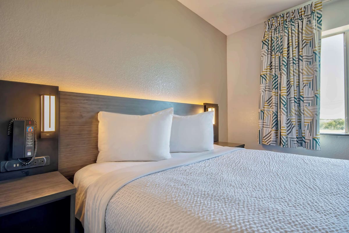 A hotel room with a bed, a bedside table with a phone, and a window with patterned curtains.