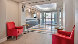 Un hall de hotel con una mesa de recepción, dos sillones rojos, piso a rayas y un tragaluz.