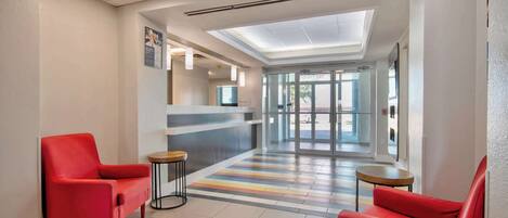 Un hall de hotel con una mesa de recepción, dos sillones rojos, piso a rayas y un tragaluz.