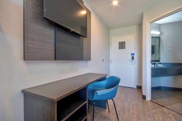 Una habitación de hotel moderna con televisor de pantalla plana en la pared, un escritorio de madera, una silla azul y un baño visible con lavamanos y espejo.