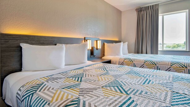 Habitación de hotel con dos camas, cabecero con luces empotradas en la pared, ventana grande con cortinas y colcha estampada.