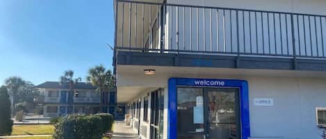 Entrada de un edificio con un letrero azul que dice 'welcome', un balcón y una señal que indica una oficina.