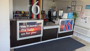 A hotel reception desk with a large number '6' sign, a sign stating 'We'll leave the light on for you.', and various amenities like snacks and a refrigerator. - Motel 6 Gulfport, MS – Airport (Gulfport)