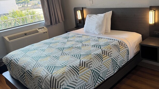 A modern hotel room with a large bed, a bedside table with a lamp, a window with curtains, and a view of greenery outside. - Motel 6 Gulfport, MS – Airport (Gulfport)