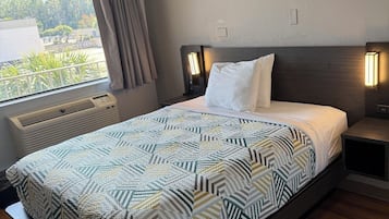 A modern hotel room with a large bed, a window with curtains, a bedside table with a lamp, and a built-in heater.