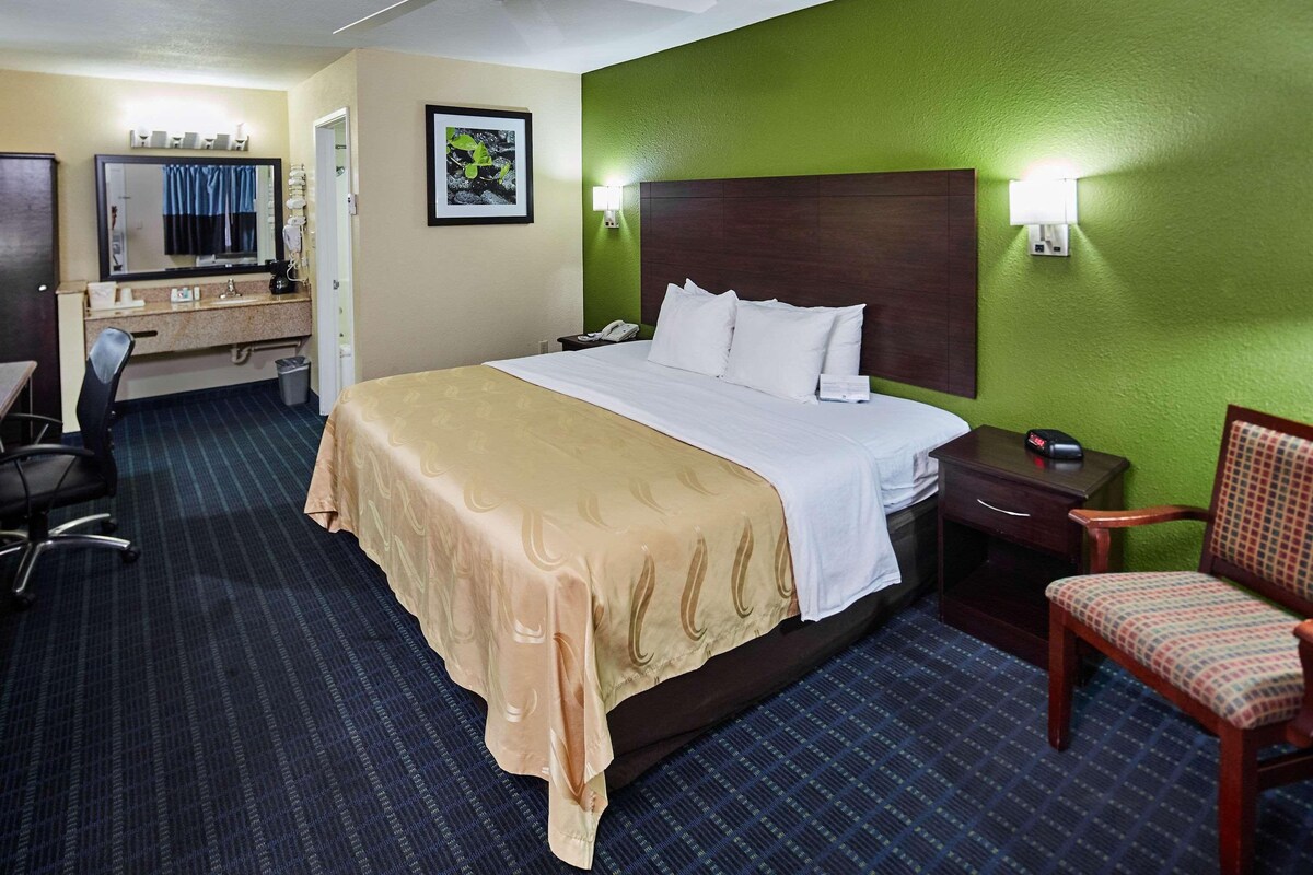 A hotel room with a bed, a desk, a chair, a nightstand, and a wall-mounted lamp.