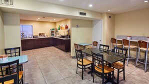 A spacious dining area with a central table surrounded by chairs, a kitchen area with a counter and appliances, and a door leading to another room.