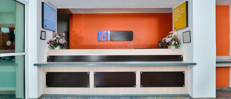 A hotel reception desk with a blue carpet marked with the number 6, a sign indicating the hotel name, and a flower arrangement.