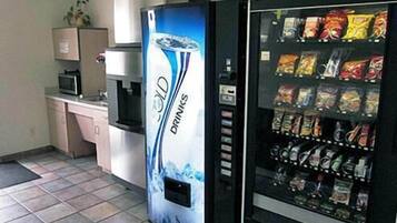 A vending machine with a drink dispenser and a snack vending machine in a room with tiled flooring and a microwave on a counter.