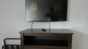A dark wooden TV stand with a flat-screen TV mounted above it, a small remote control on the stand, and a metal rack with hooks to the left. - Americas Best Value Inn & Suites Groves Port Arthur (Groves)