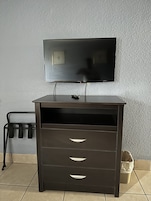 Un mueble de televisión de madera oscura con un televisor de pantalla plana montado encima, un control remoto pequeño sobre el mueble y una estructura metálica con ganchos a la izquierda.