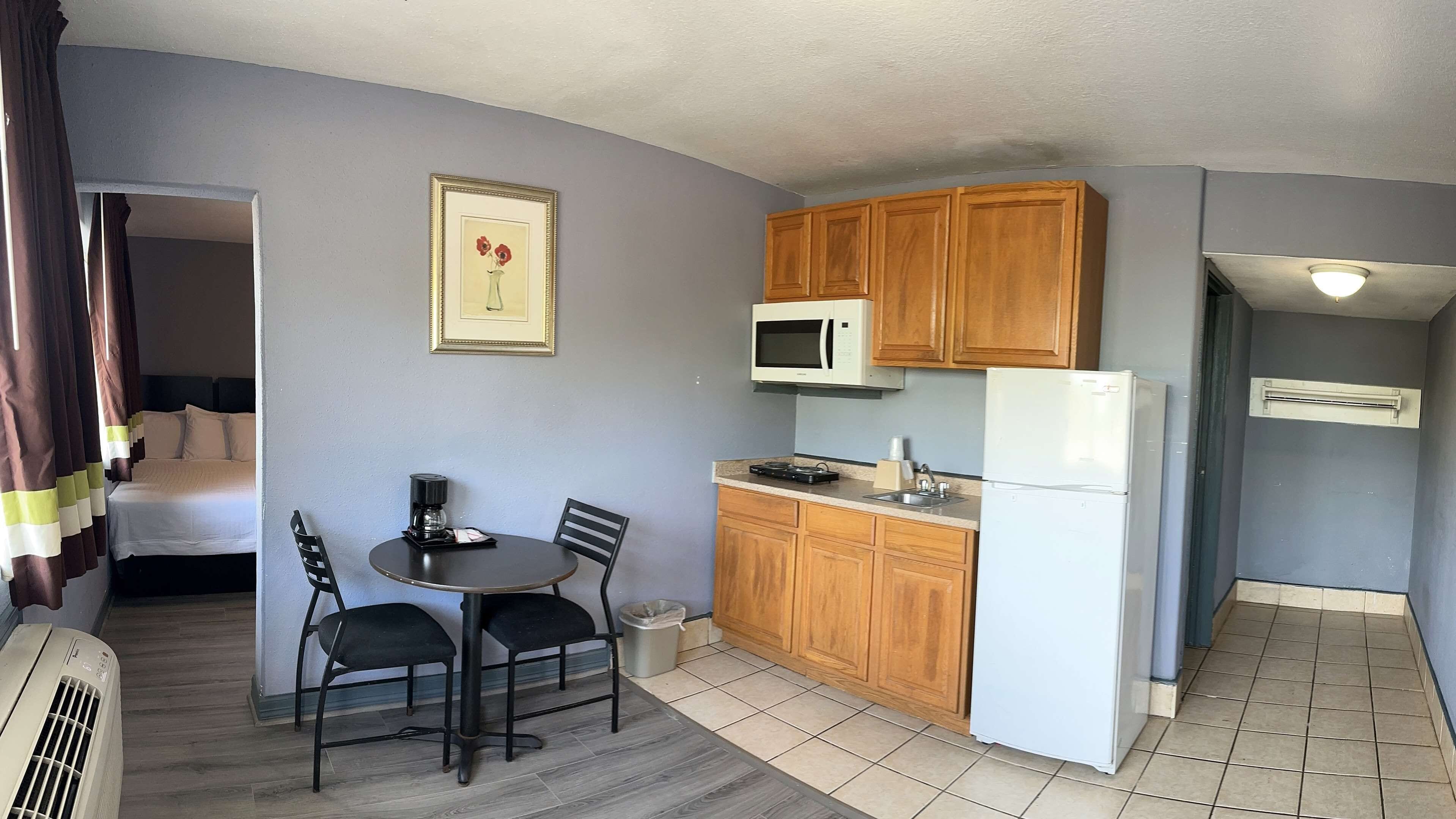 A compact hotel room with a kitchenette, a bed, a dining area with two chairs, and a framed picture on the wall.