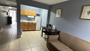 A living room with a beige sofa, a dining table with chairs, and a kitchen area with wooden cabinets and a refrigerator. - Americas Best Value Inn & Suites Groves Port Arthur (Groves)