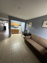 Una sala de estar con un sofá color beige, una mesa de comedor con sillas y una zona de cocina con armarios de madera y un refrigerador.