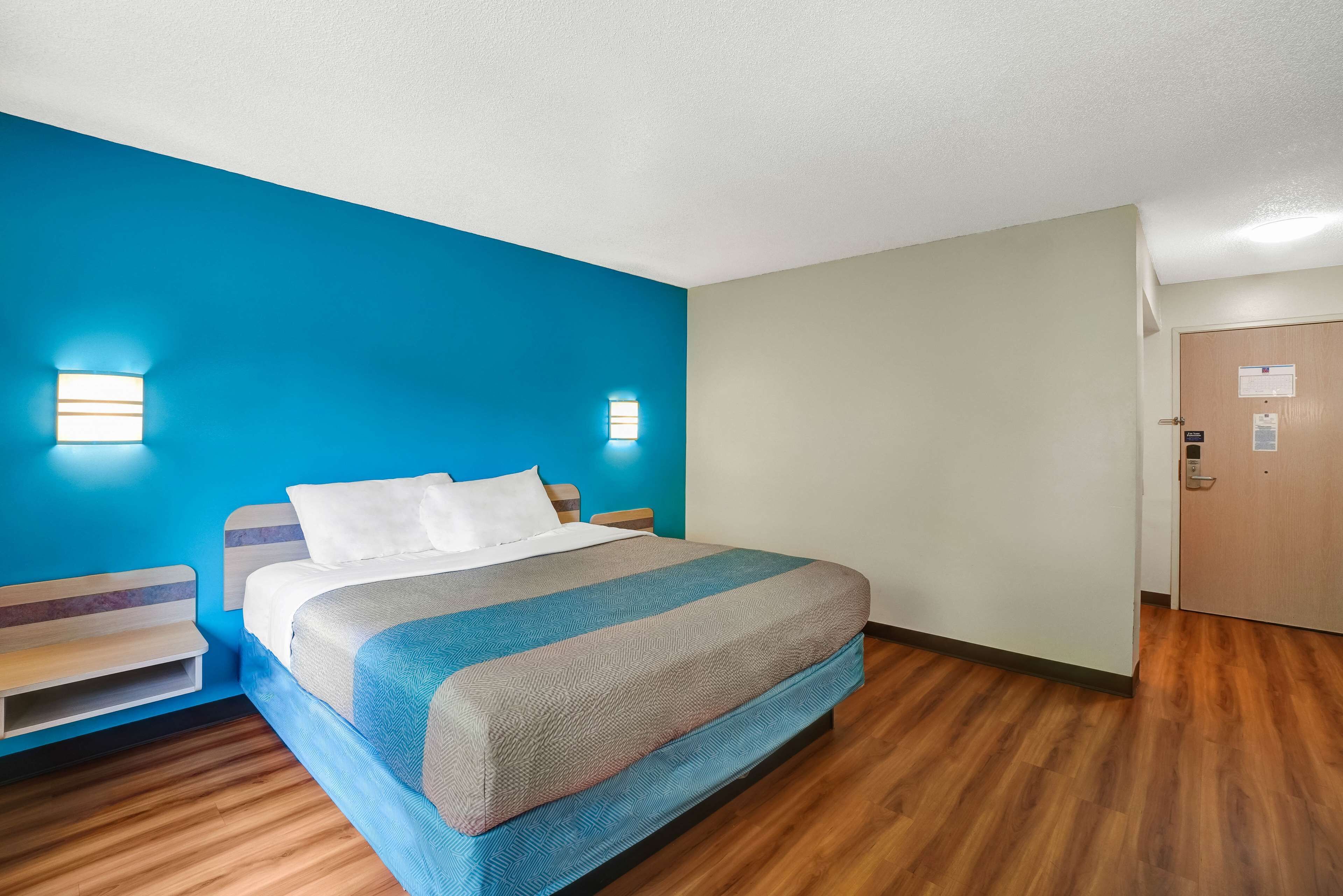 Une chambre d’hôtel avec un lit, une table de chevet, une étagère fixée au mur et une porte.