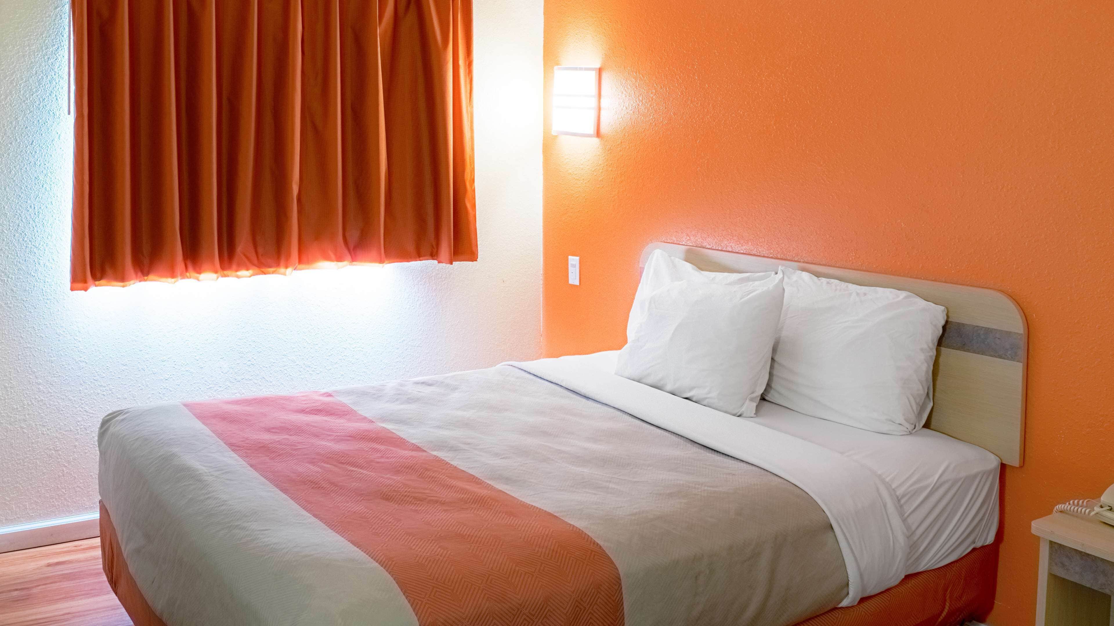 A hotel room with a bed, orange walls, a window with curtains, and a bedside table with a phone.
