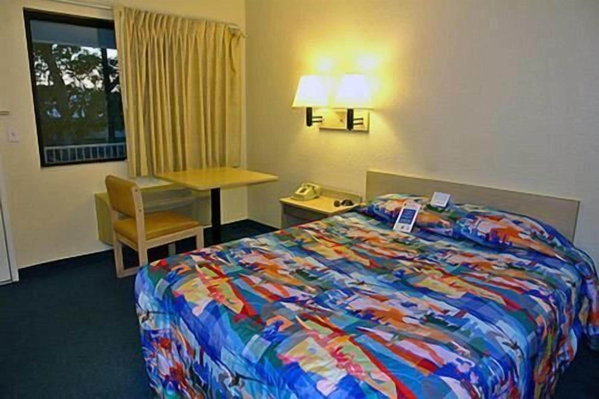 A hotel room with a bed, a desk, a chair, a nightstand, and a window with curtains.