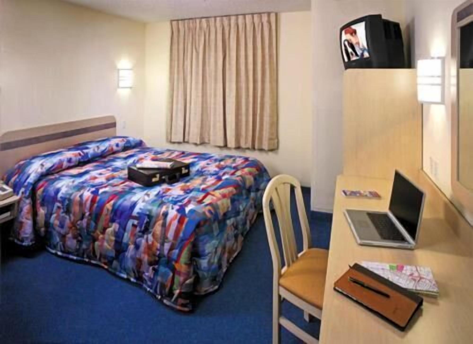 A hotel room with a bed, a desk with a laptop, a chair, a television, and a window with curtains.