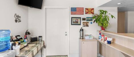 A small kitchenette with a coffee maker, a water dispenser, and a coffee machine. There is a TV mounted on the wall and a plant on the counter.