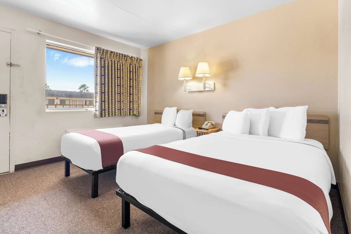 Une chambre d’hôtel avec deux lits, une fenêtre avec des rideaux, une lampe fixée au mur et une porte.