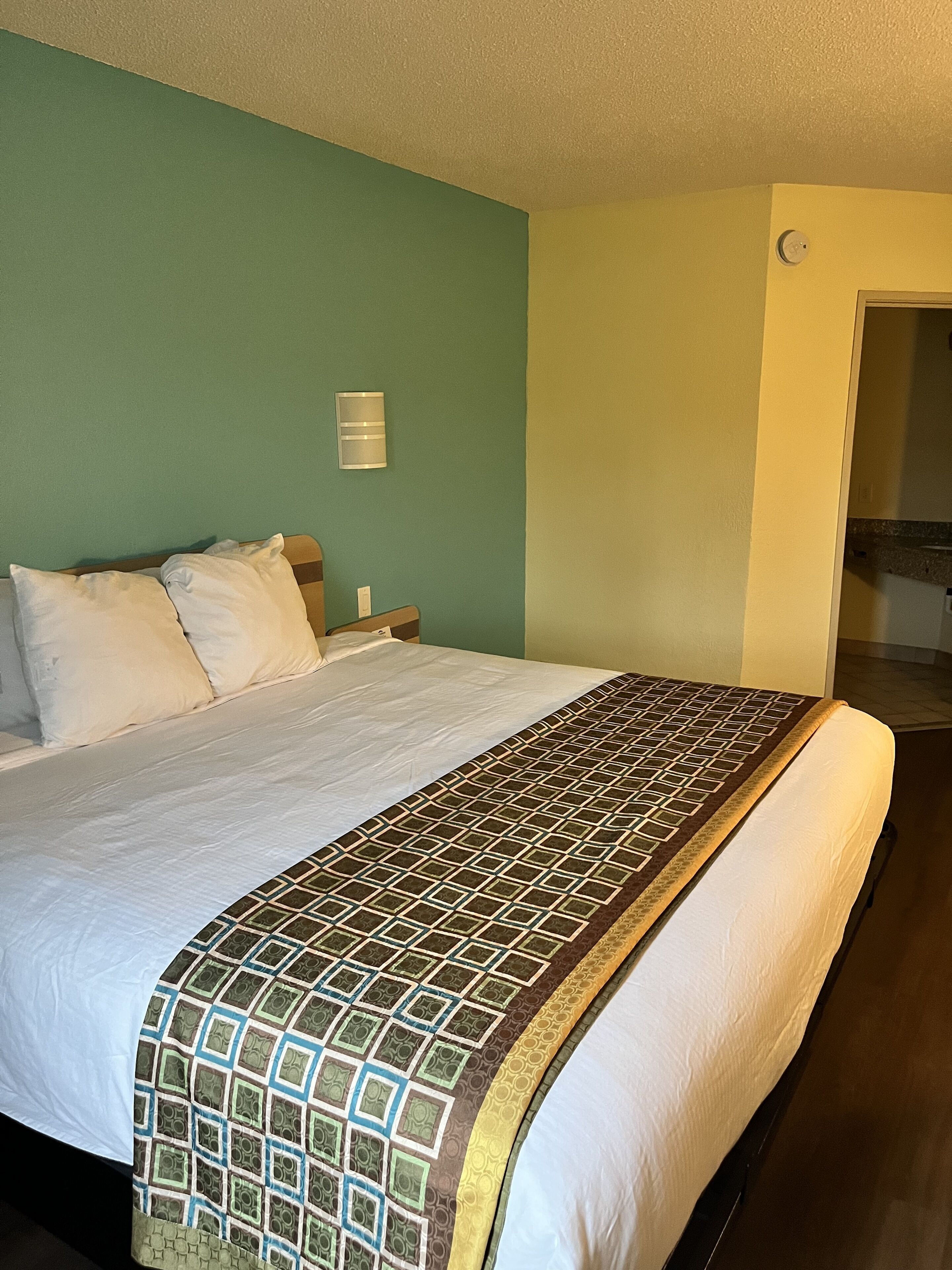 Ein Hotelzimmer mit Bett, Nachttisch, Wanduhr und einer Tür zu einem anderen Raum.