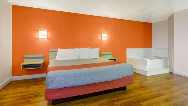 A bedroom with a large bed, two bedside tables, a nightstand, a built-in bathtub, and orange walls. - Motel 6 Beaver, UT (Beaver)