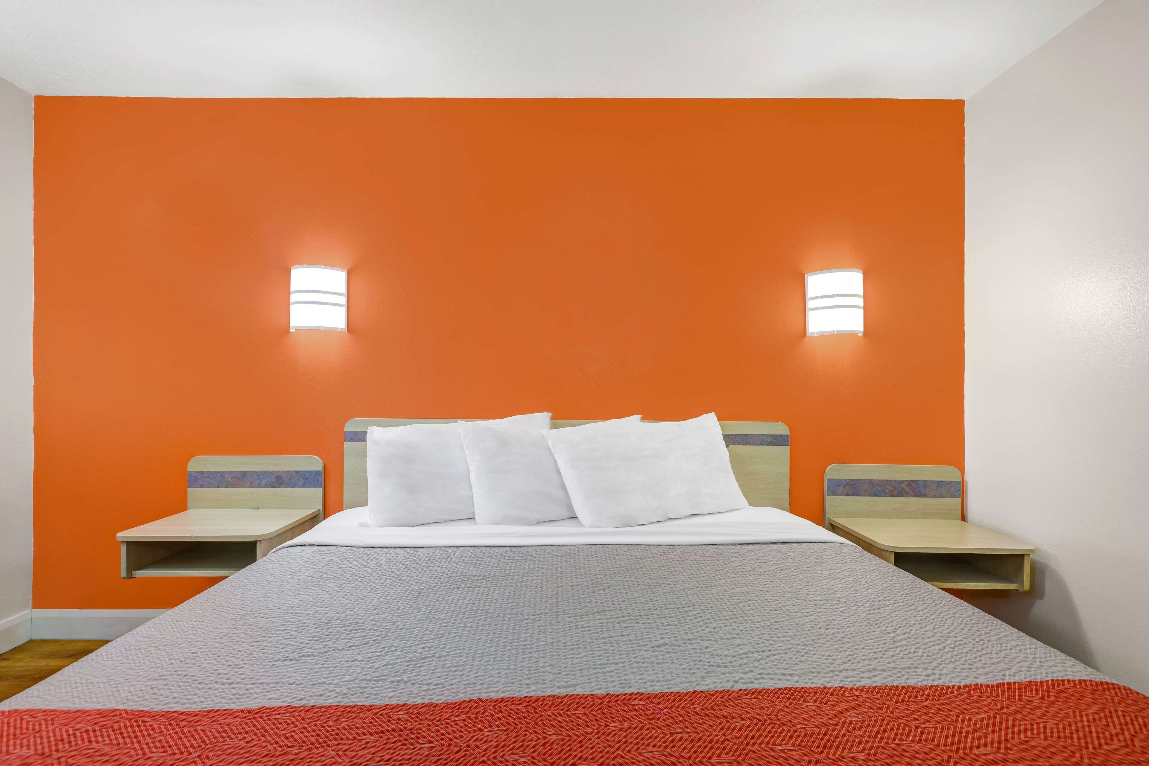 Hotellihuone, jossa on oranssi tehosteseinä, suuri sänky valkoisilla vuodevaatteilla, kaksi yöpöytää ja kaksi seinään kiinnitettyä valaisinta.