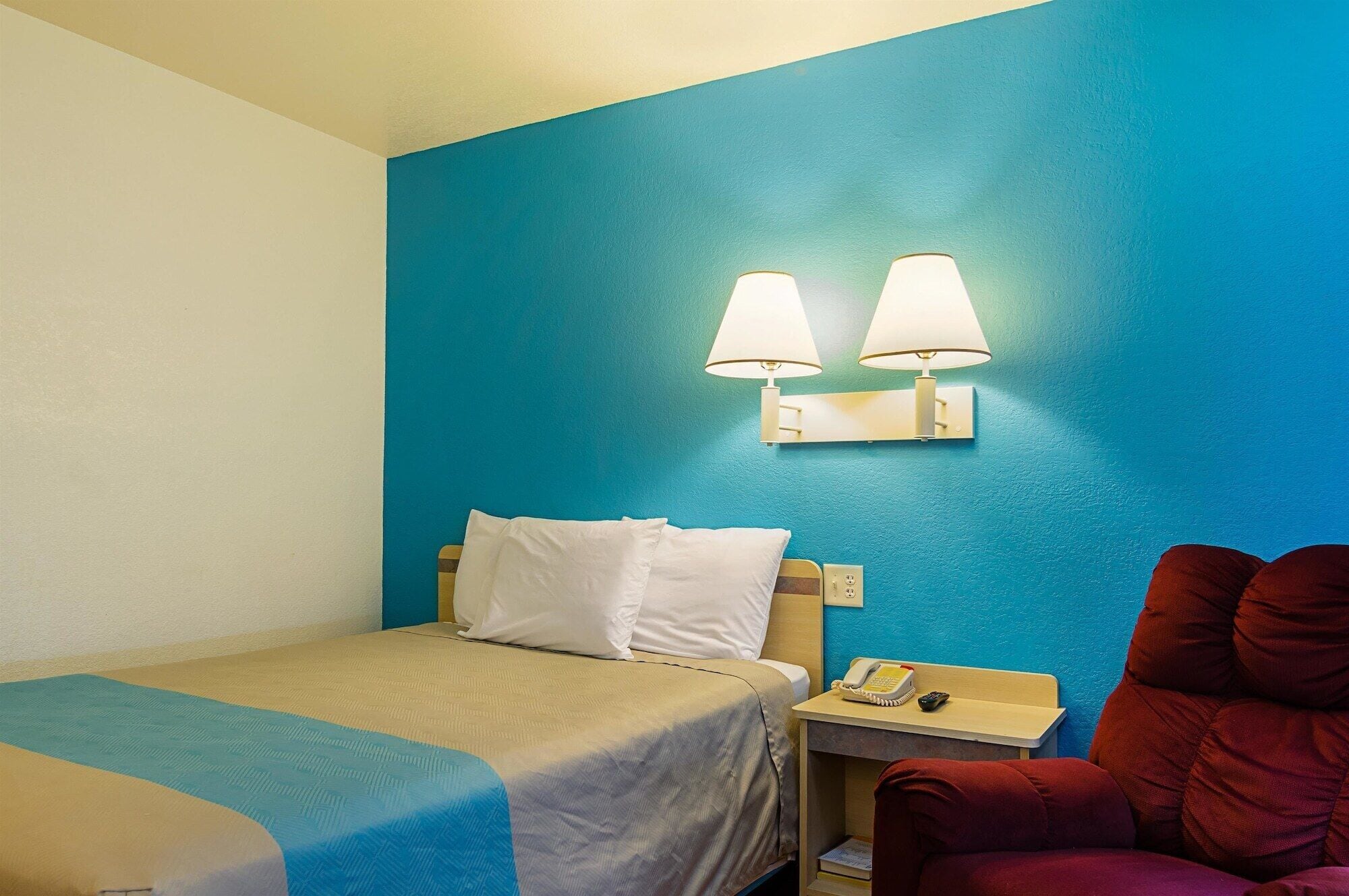 Habitación de hotel con una cama, una mesita de noche con lámpara, una silla roja y una pared decorativa azul.