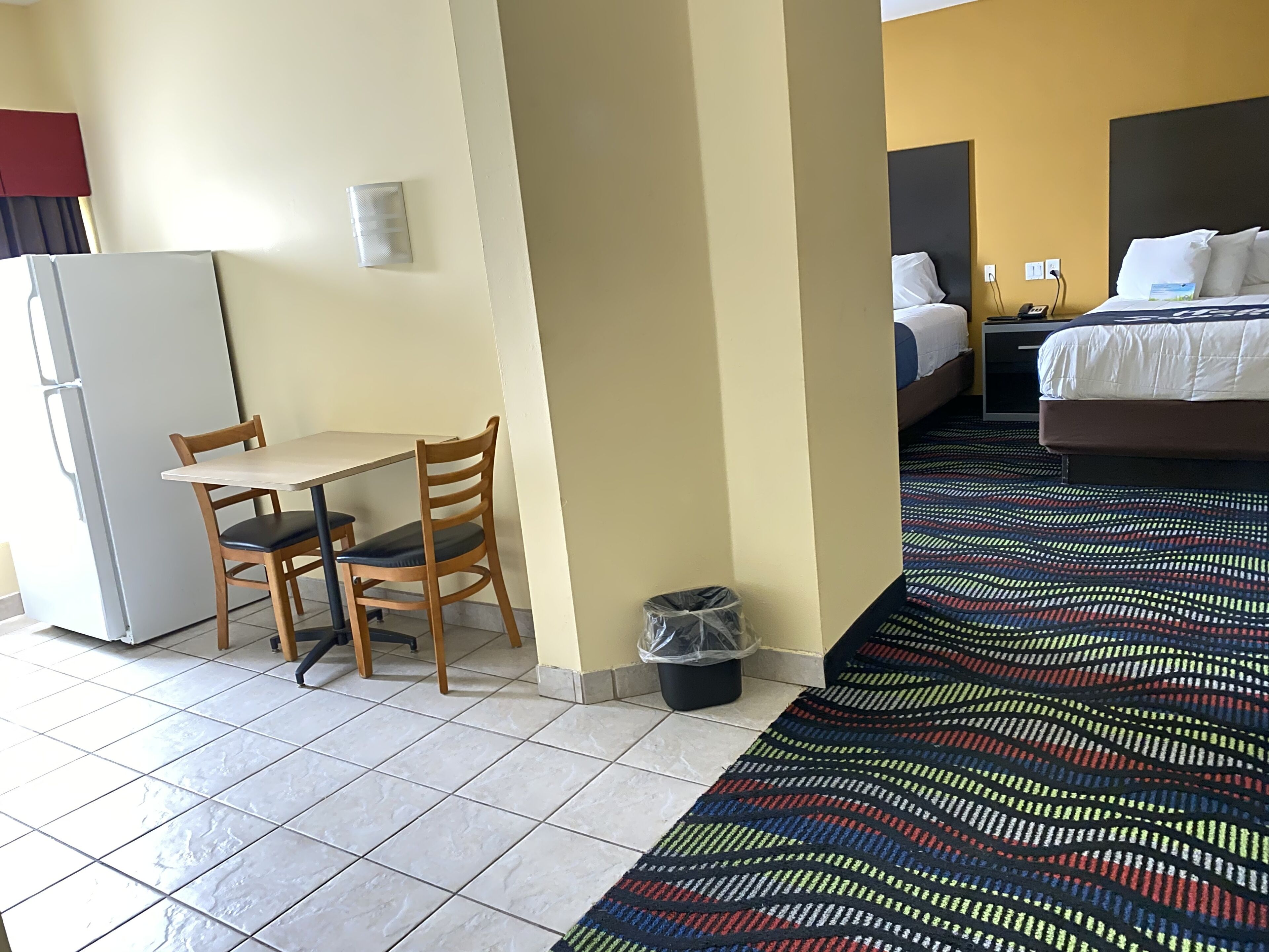 Una habitación de hotel con cama, escritorio con silla, un cesto de basura y una alfombra estampada.